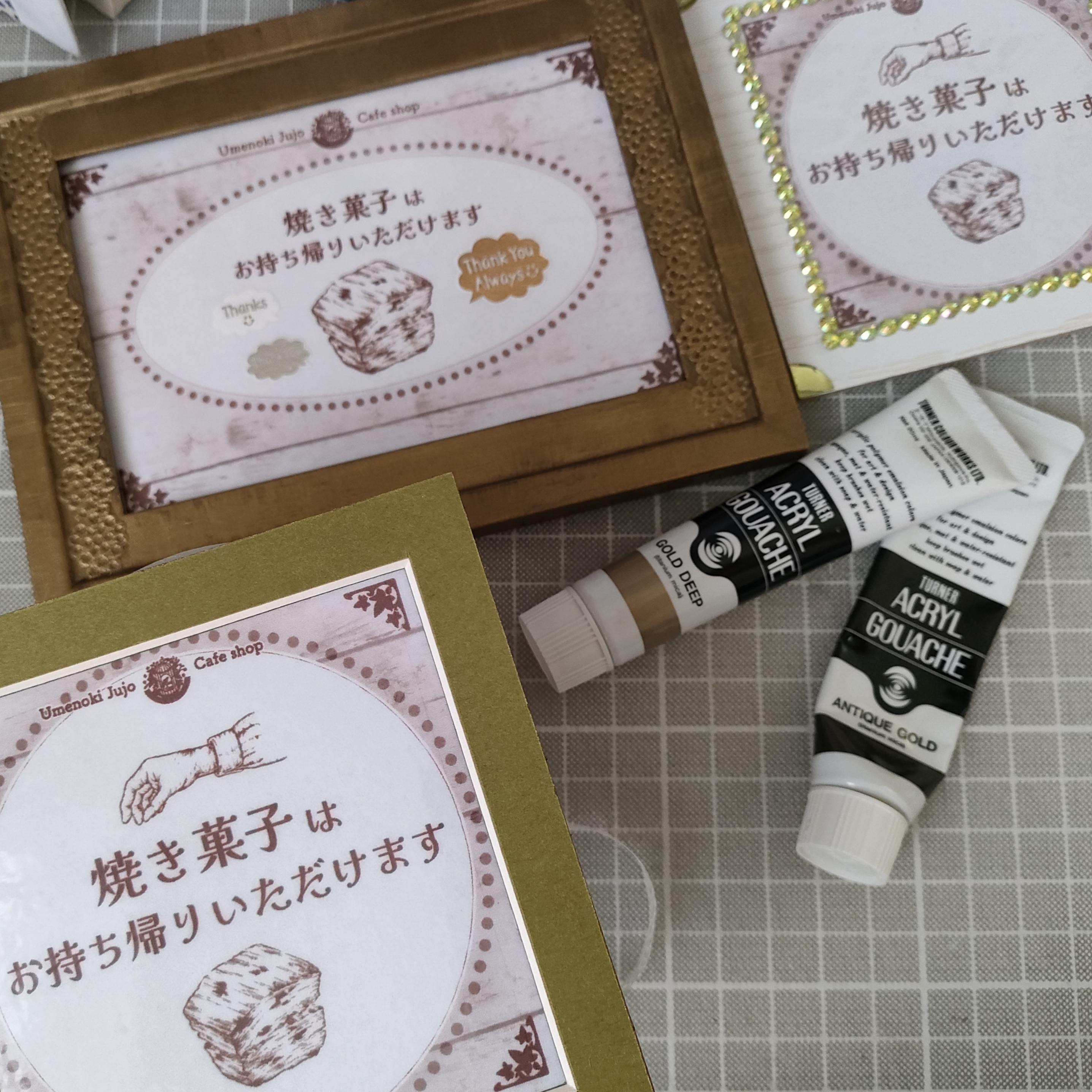 梅の木十条店さんの、焼き菓子お持ちかり可を宣伝するミニ看板制作風景です。オリーブグリーンのマット紙をフレームにしたものや、100円均一のフォトフレームを金色のアクリル絵の具で塗りアンティーク感を出したものなど、3種類のフレームに、スコーンや手を描いたペン画イラストとテキストをレイアウトしています。