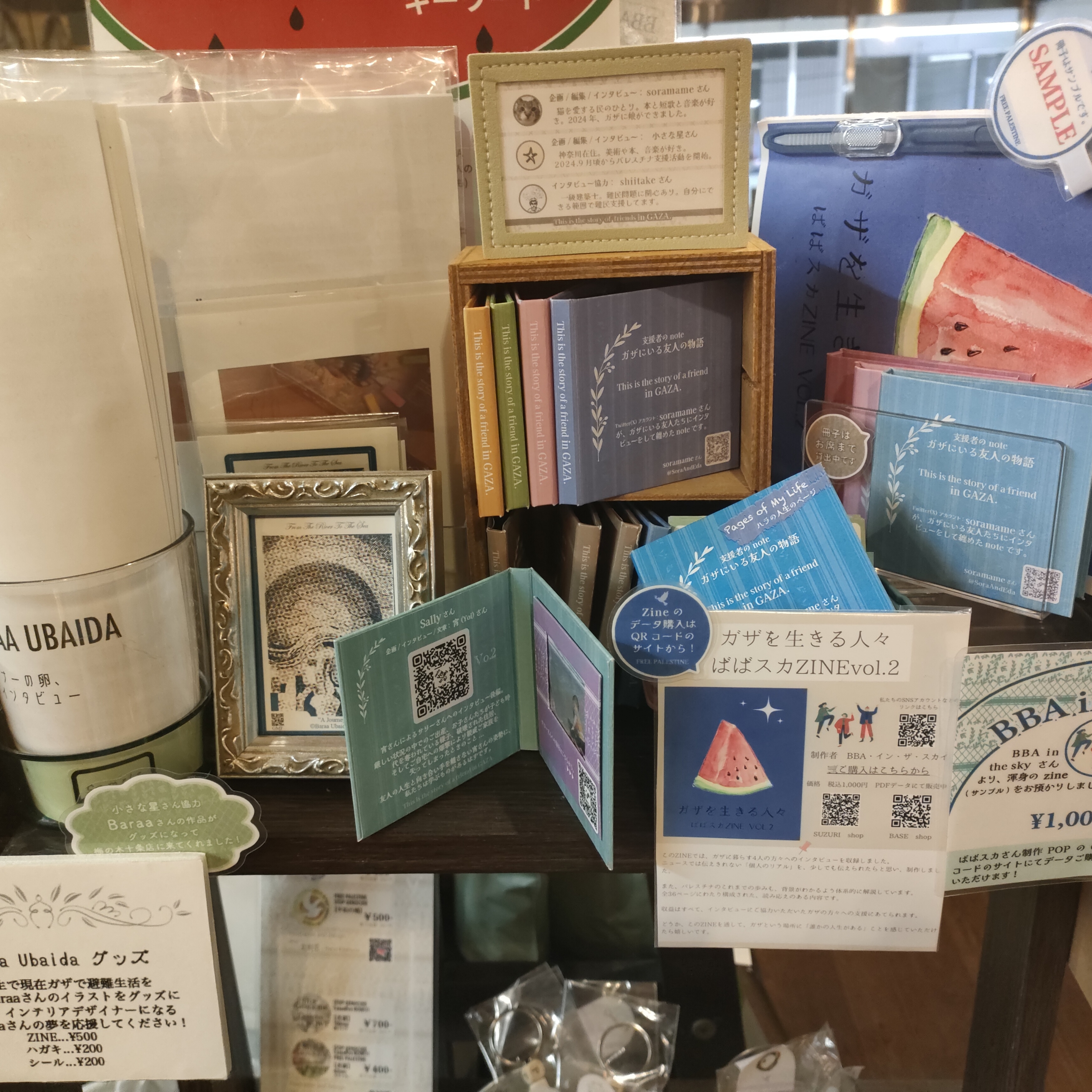 梅の木十条店さんレジ横の物販スペースをお借りして展開している支援グッズ販売棚の様子や、支援者のnoteを紹介するミニ冊子を撮影した画像です。
