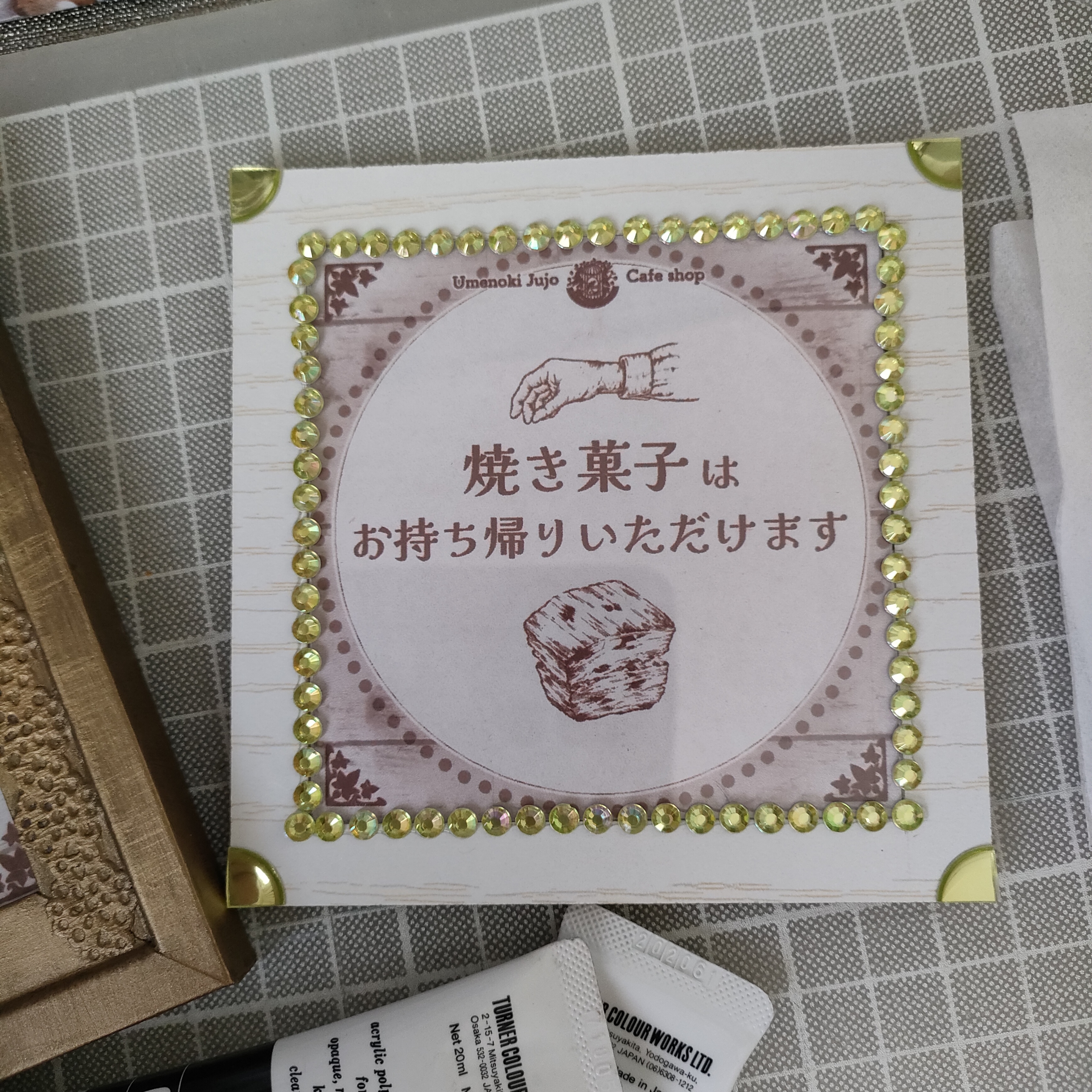 梅の木十条店さんの、焼き菓子お持ちかり可を宣伝するミニ看板制作風景です。白い木目フレームの縁にイエローグリーンのラインストーンシールを飾り、スコーンや手を描いたペン画イラストとテキストをレイアウトしています。